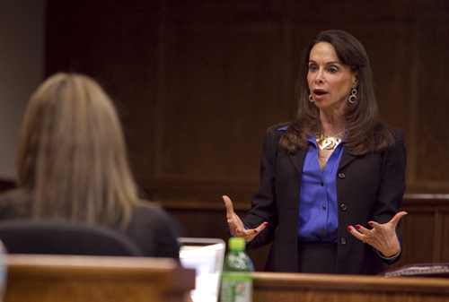 A dark-haired woman wearing a black blazer and purple shirt speaks in front of a practice courtroom.
