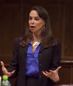 A woman with long dark hair, a blue shirt, and a dark suit jacket speaks to a student in a practice courtroom.