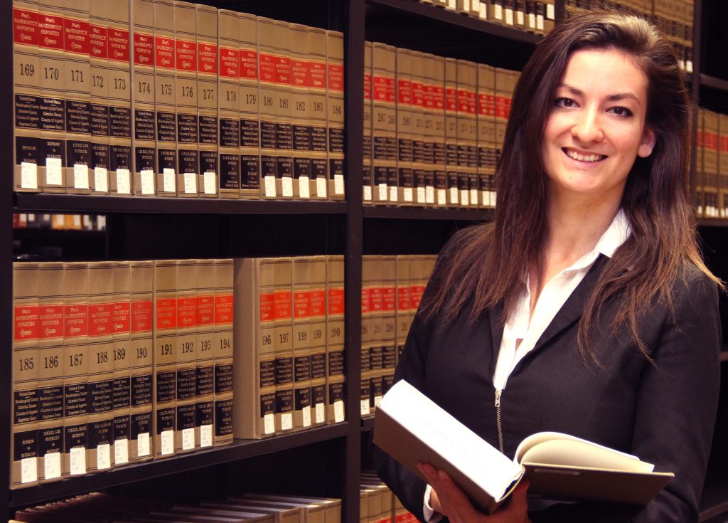 Alumna Viviana Vasiu is dressed in a suit and standing in front of a large library shelf full of law books.
