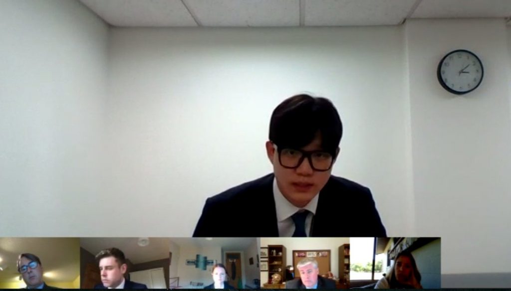 A screen shot of a virtual competition round in which a young man with dark hair argues his case on camera. Judges are featured in smaller frames below him.