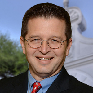 A headshot of a male law professor smiling and wearing glasses outdoors.