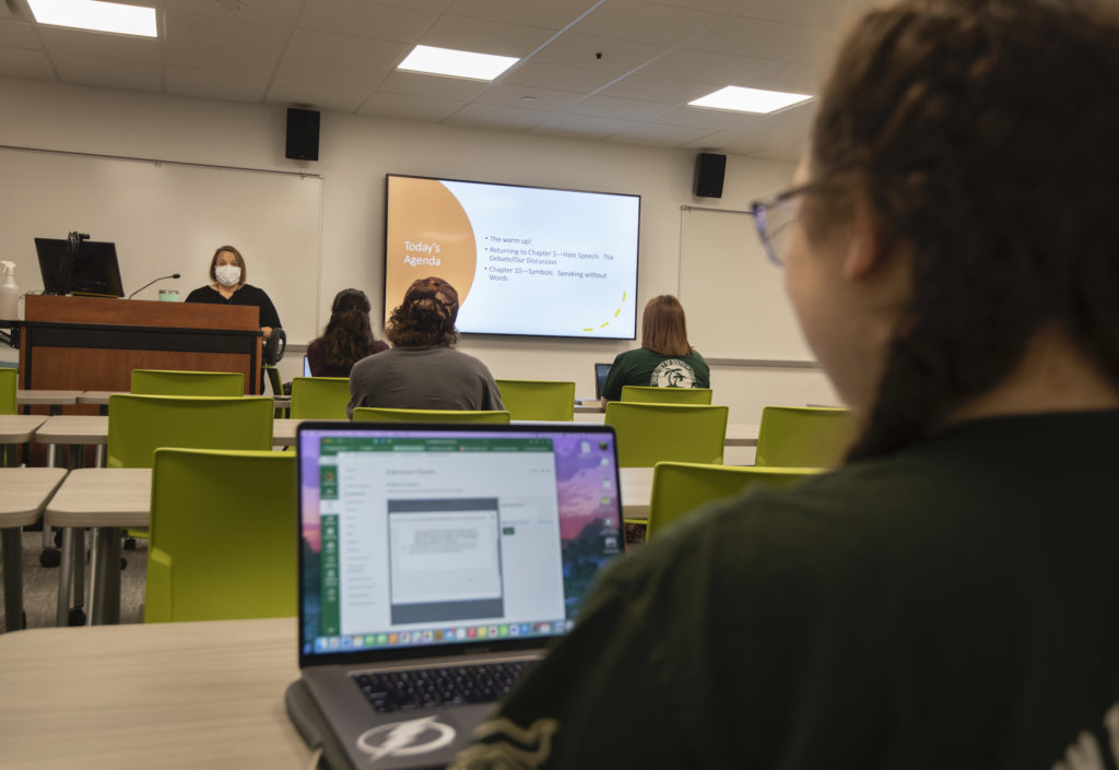 A student at the back of the room sits at a laptop while a professor gives a lecture