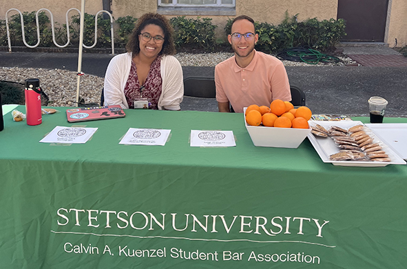 Two students sit at a table featuring promotional materials for the Calvin A. Kuenzel Student Bar Association, a bowl of oranges, and a plate of cookies.