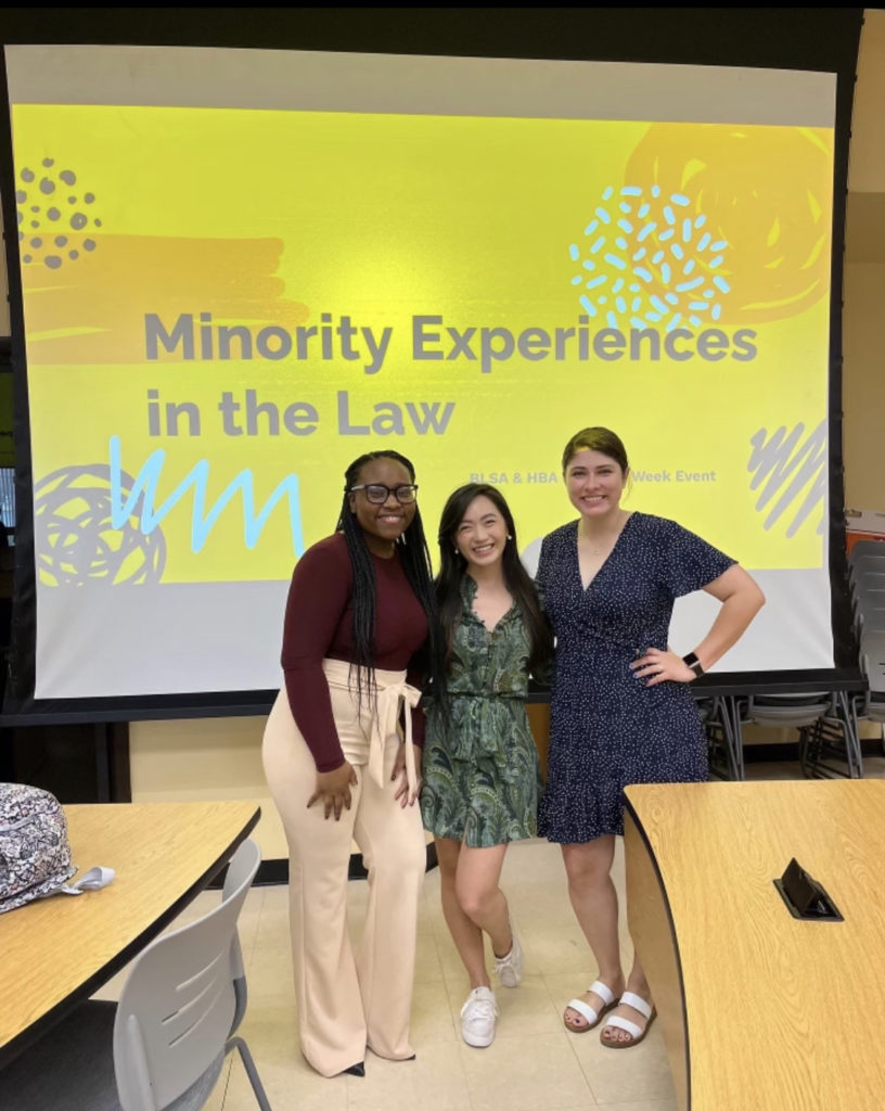 Three young women stand in front of a presentation slide that reads "Minority Experiences in the Law"