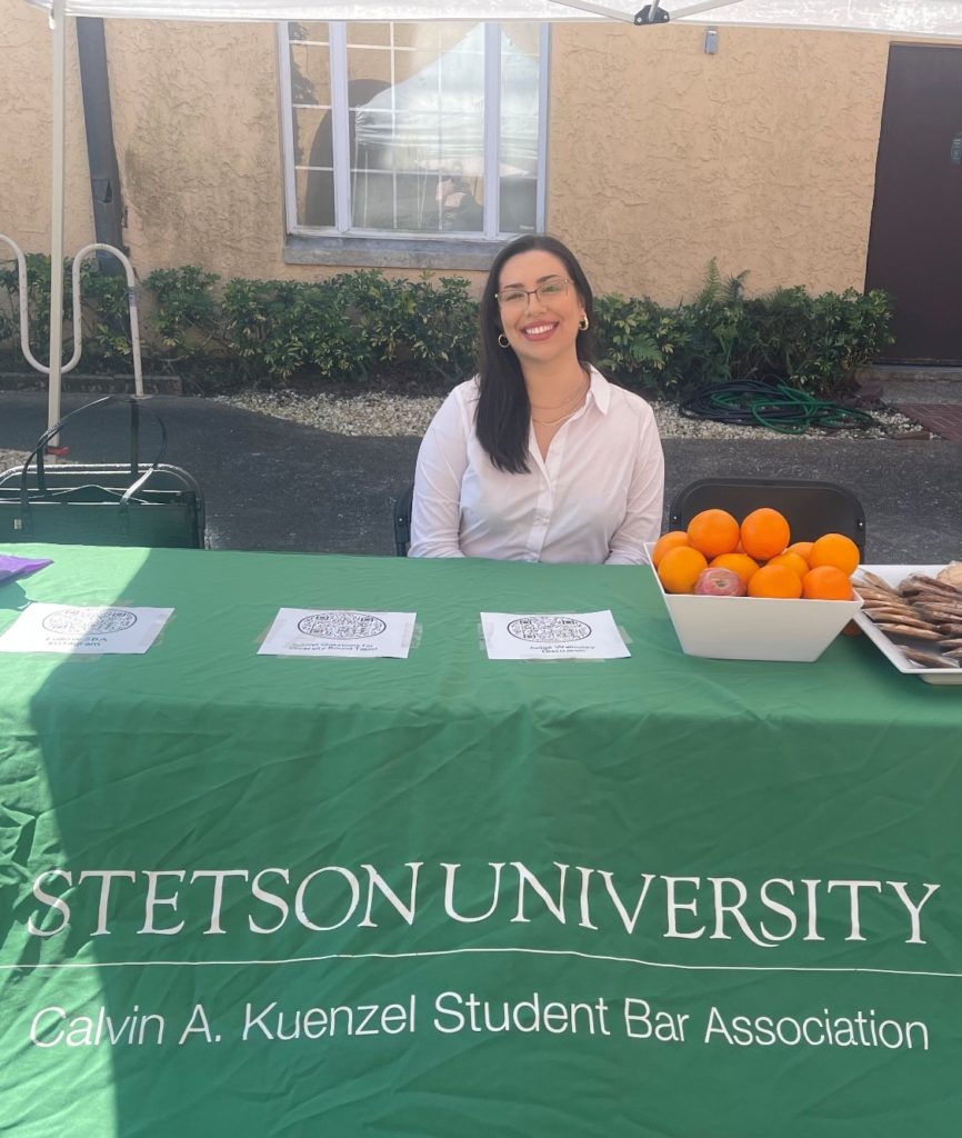 A law student sits at a table with a bowl of oranges and a plate of cookies. A tablecloth reads "Stetson University Calvin A. Kuenzel Student Bar Association."