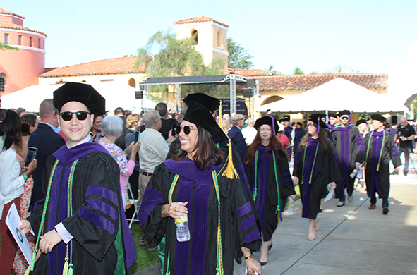 Graduates in sunglasses smile as they leave an outdoor commencement ceremony