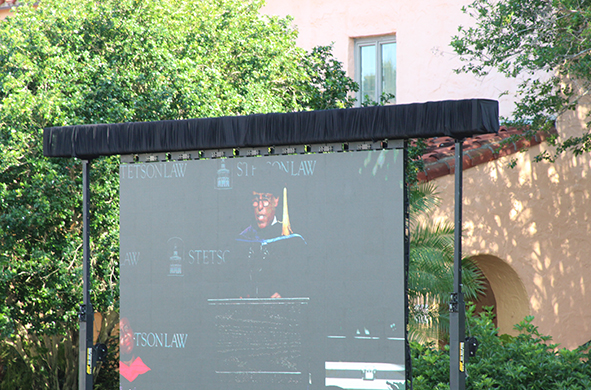 A speaker is seen on a large screen at graduation