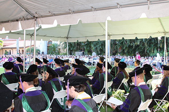 Students in graduation caps and gowns wait under a canopy.