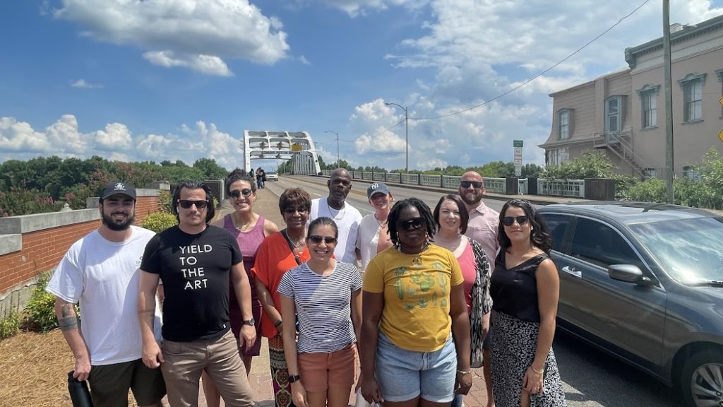 Eleven people stand on the side of the road near a bridge on a sunny day.