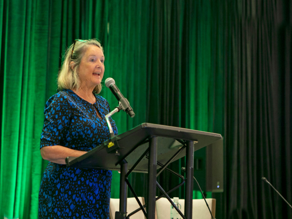A blonde female professor in a blue and black dress speaks at a podium in front of a green curtain.