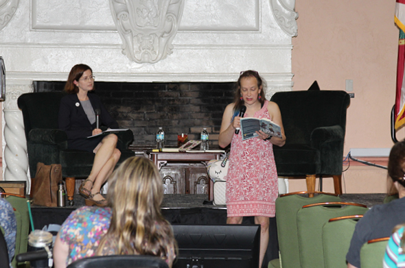 Professor Kristen Adams and Dr. Stephanie Burt sit on a stage in the Great Hall; Dr. Burt is reading from a book of her poetry.