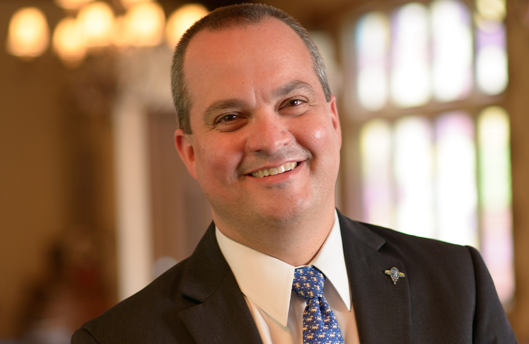 A closeup shot of a man smiling and wearing a black suit jacket and blue tie.