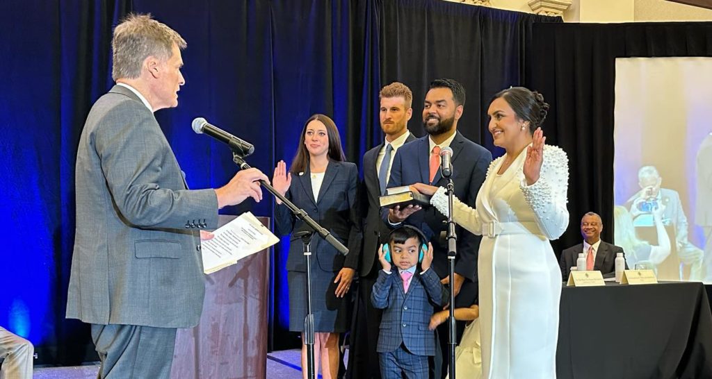 A woman smiles and raises her left hand in the air while being sworn into office on a book. Her family stands next to her smiling.