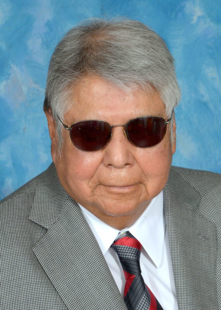 A headshot of a man with grey hair wearing sunglasses and a grey suit with a colorful tie.