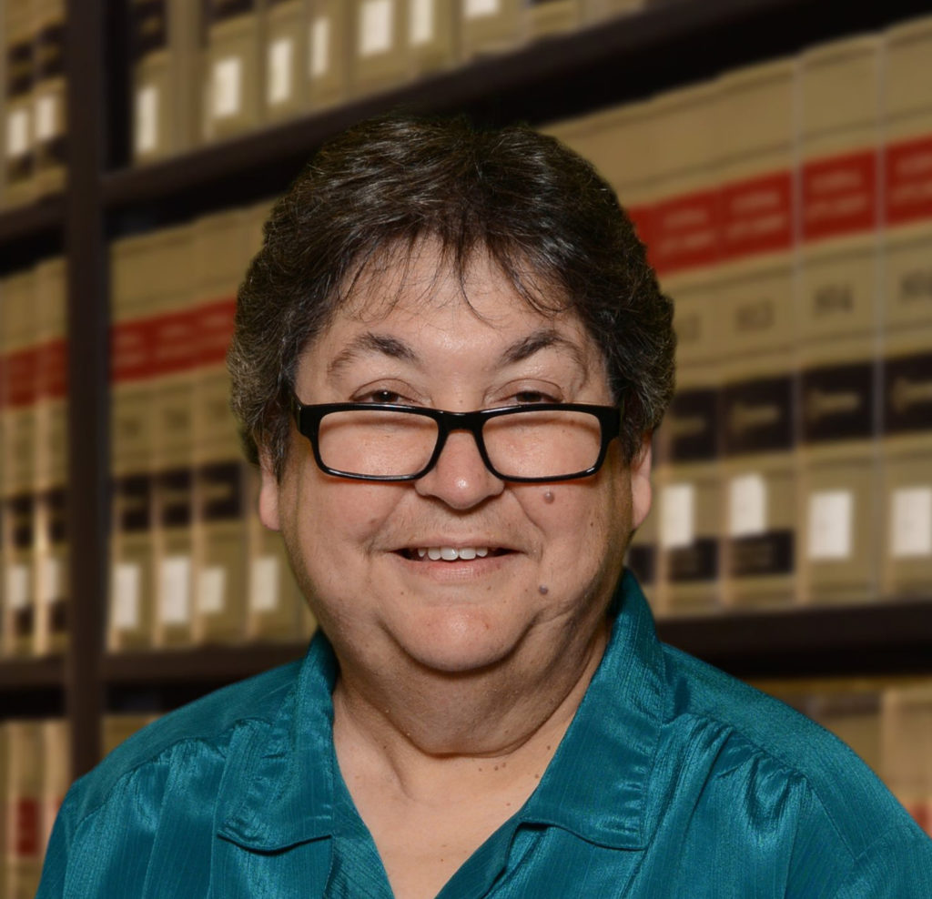A headshot of a woman wearing a green paisley shirt and glasses standing in front of a shelf full of law books.