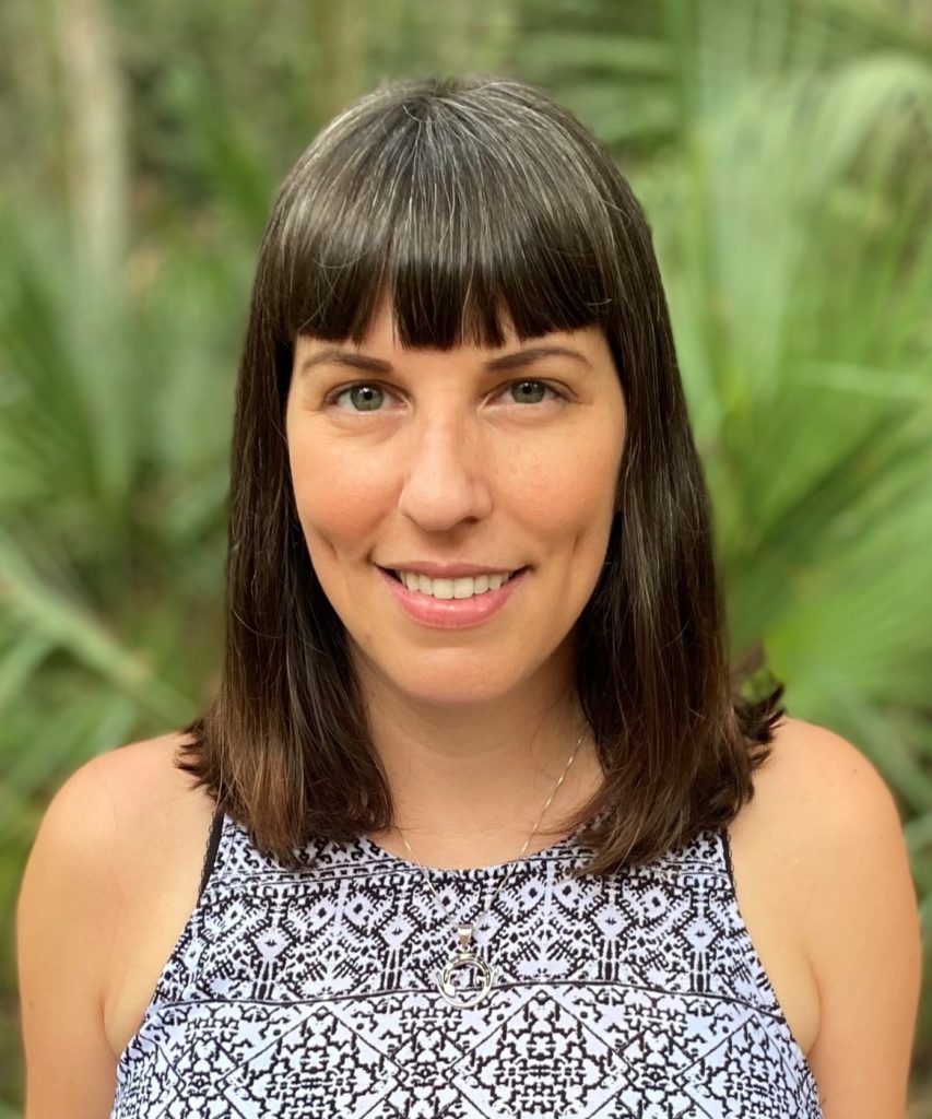 A headshot of a young Caucasian brunette against a backdrop of greenery.