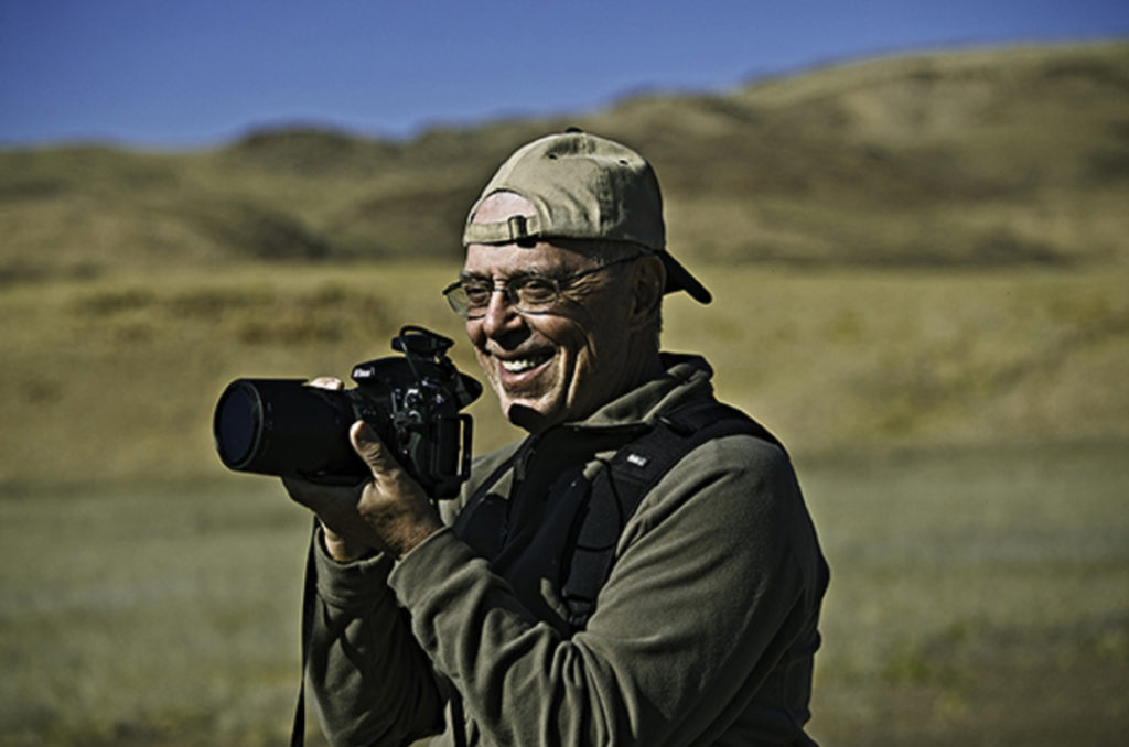 A man with a backwards hat holds a camera on a grassy hillside in Namibia.