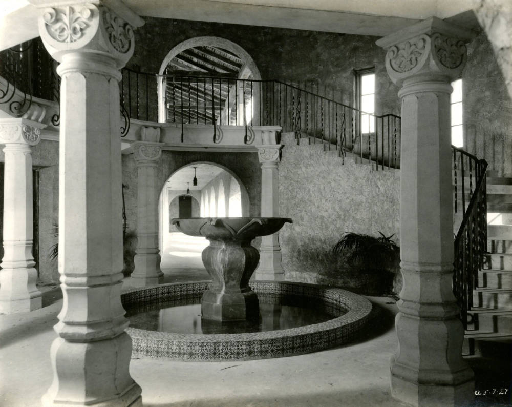 A black-and-white image of a large fountain in a circular room lined by a wide spiral staircase.
