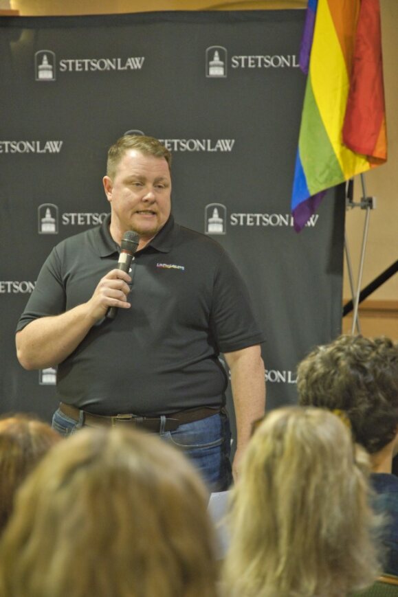 A man speaks in front of a black backdrop printed with the Stetson Law logo. A rainbow flag is in the background,