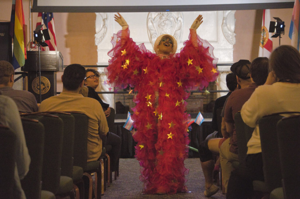 A drag performer in a voluminous, star-studded fuschia robe throws their arms in the air as they perform.