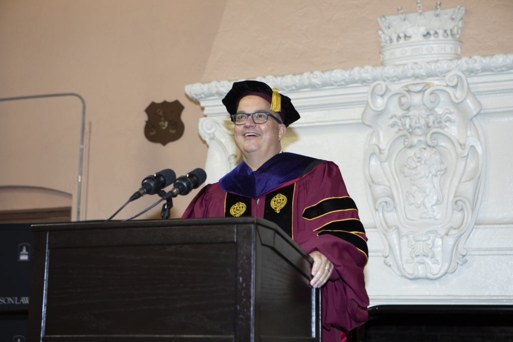 A man in faculty regalia addresses an audience from behind a podium.