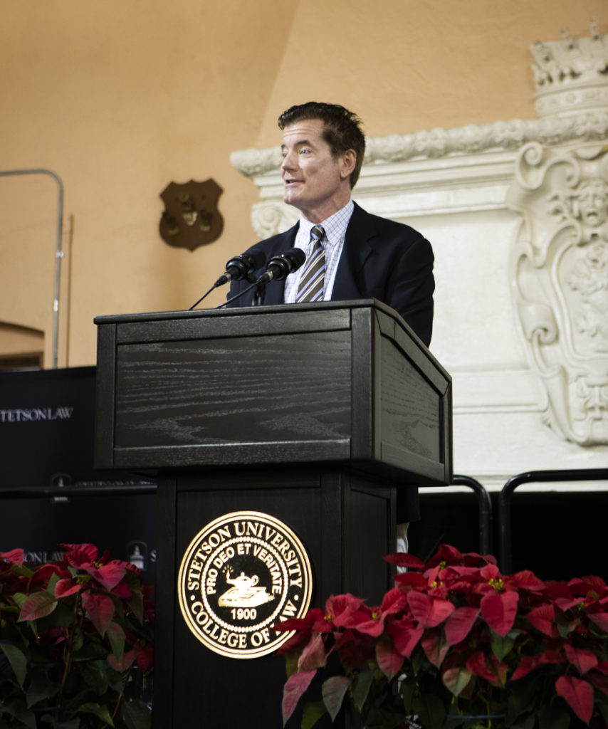 A professor speaks at a podium. Poinsettia plants are in the foreground.