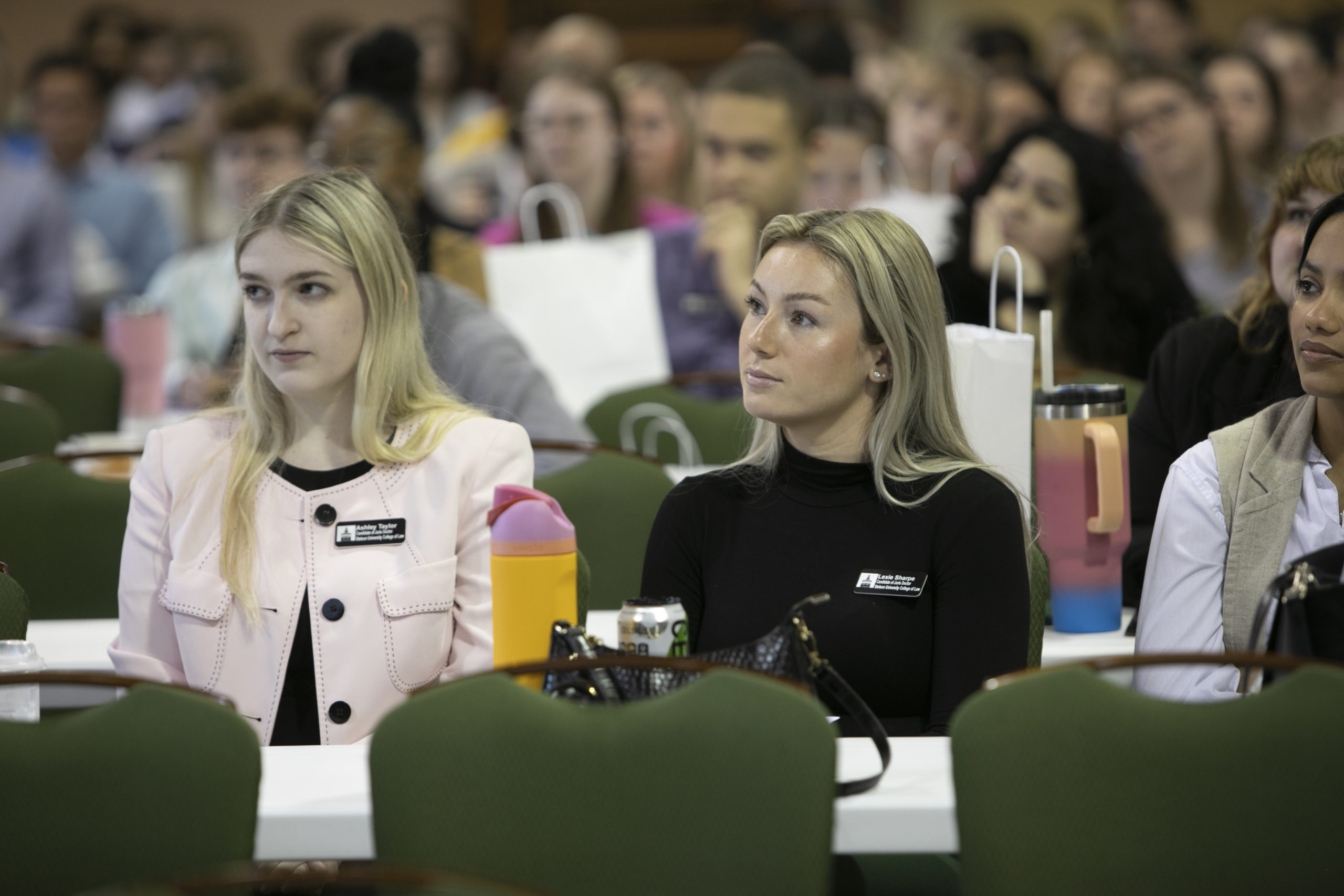 Two blonde young woman look on as a speaker addresses the audience.