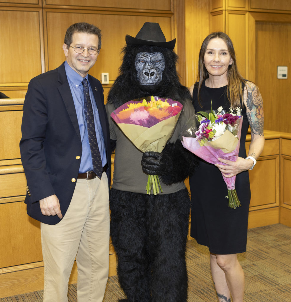 A person in a gorilla suit and two professors smile for the camera and hold bouquets of flowers.