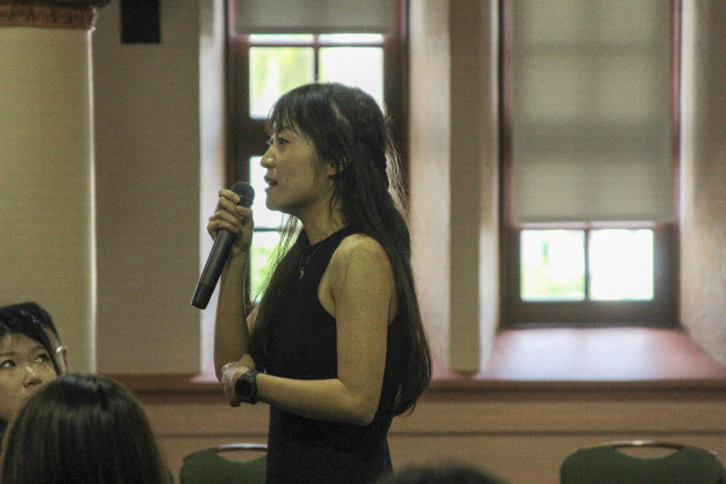 A woman with long dark hair and a sleeveless black dress stands with a microphone