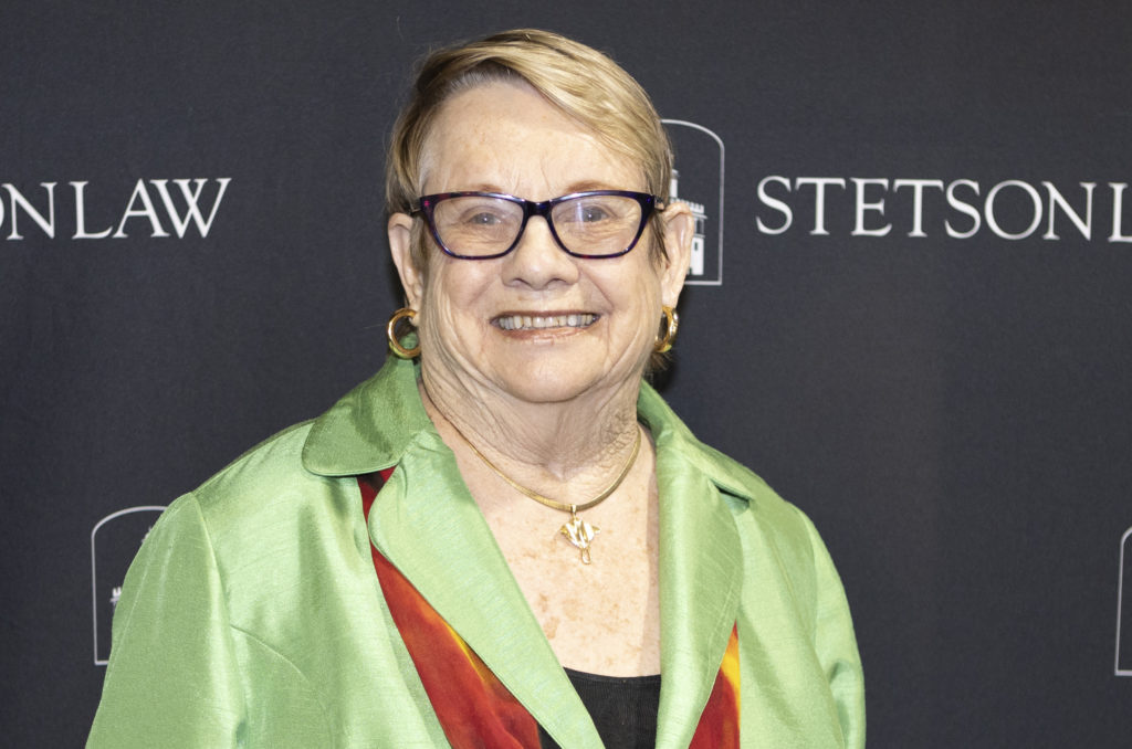 A woman with short blonde hair who wears a lime green jacket and glasses smiles at the camera.