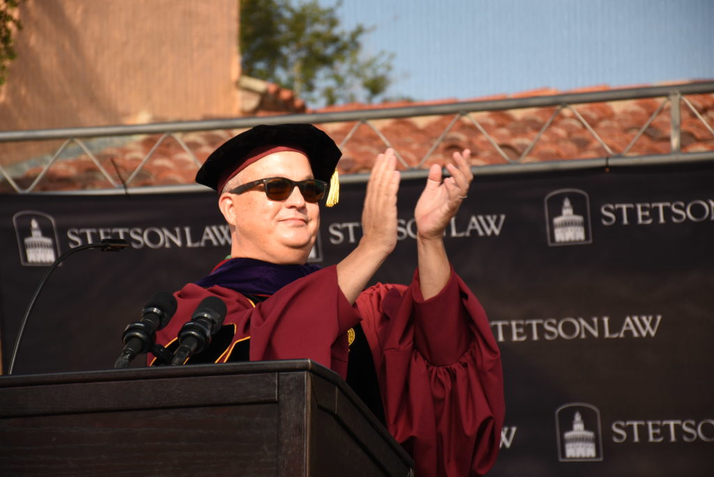 A man wearing a red ceremonial robe, a black cap, and sunglasses claps his hands behind a podium during a graduation ceremony.