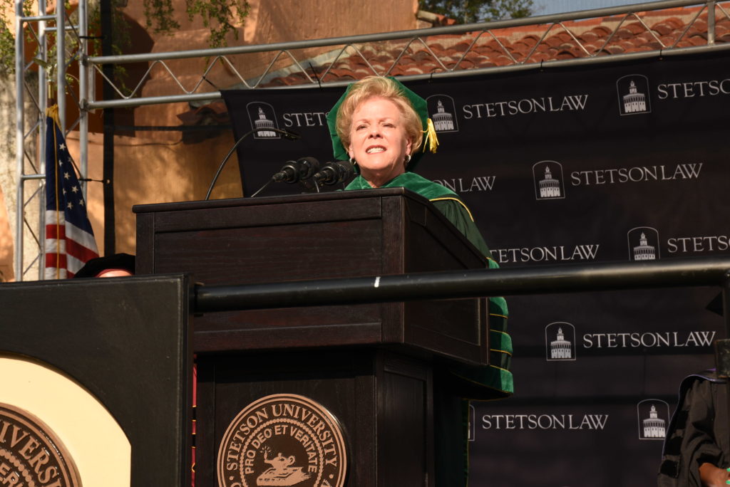 A woman with short hair wearing a green graduation ceremony robe and cap stands behind a podium and delivers a speech.