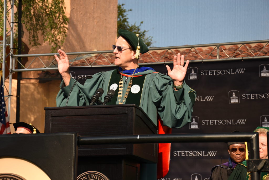 A tall man in a green robe speaks expressively at a podium