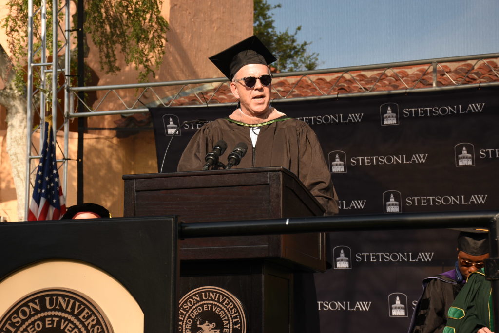 A man wearing sunglasses and a cap and gown speaks at an outdoor graduation ceremony.