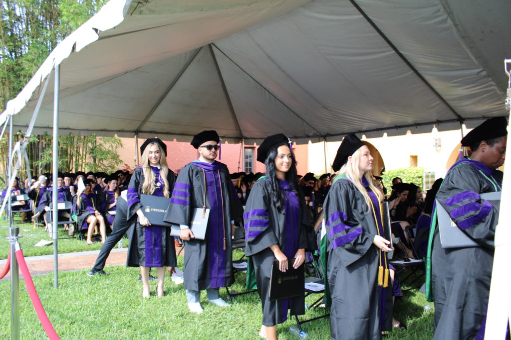 A group of students in black and purple robes and black caps stand in line waiting to walk across the stage during spring 2024 commencement.