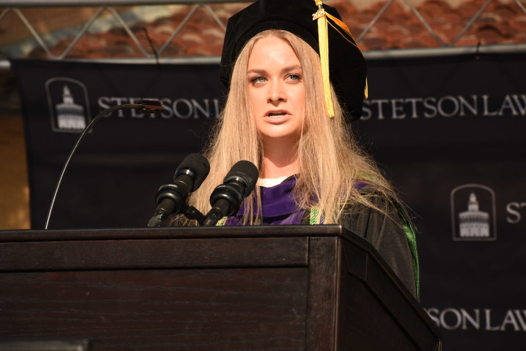 A close-up shot of a young woman with long blonde hair wearing a cap and gown as she speaks at a graduation ceremony.