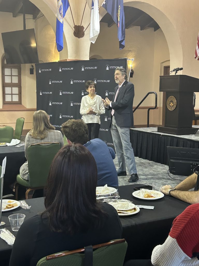A close shot of a man and a woman standing in front of a room full of students who are eating dinner.