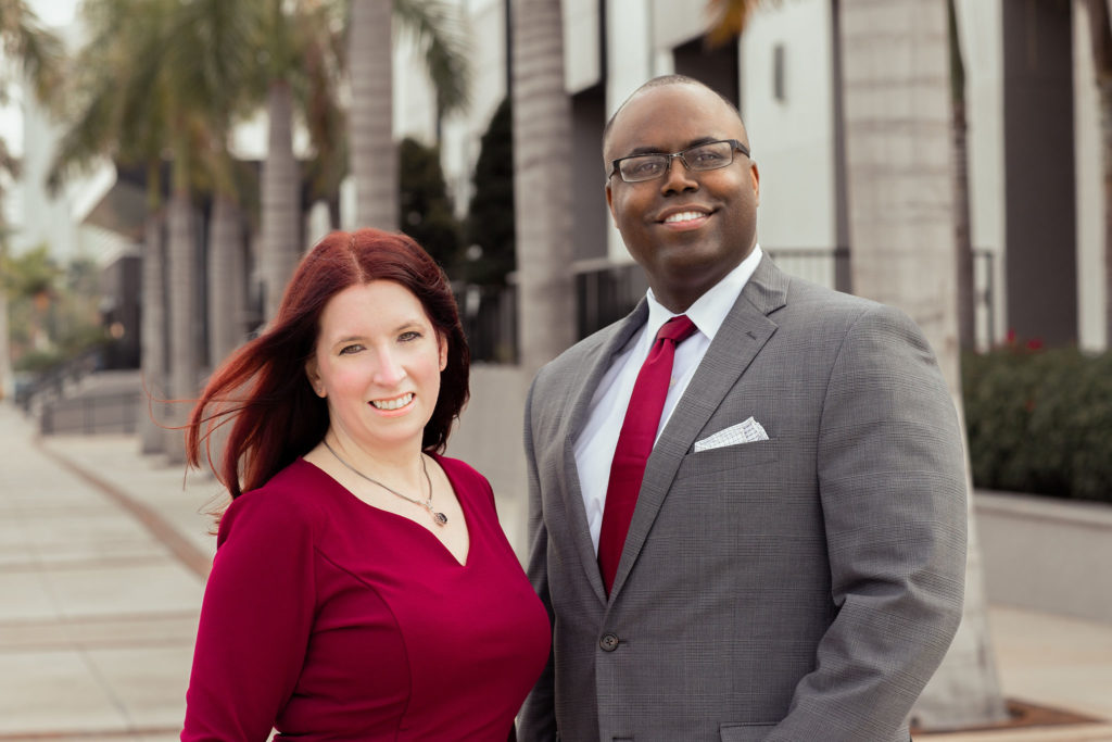 A woman with red hair wearing a red top stands next to a man with glasses wearing a grey suit jacket and red tie.