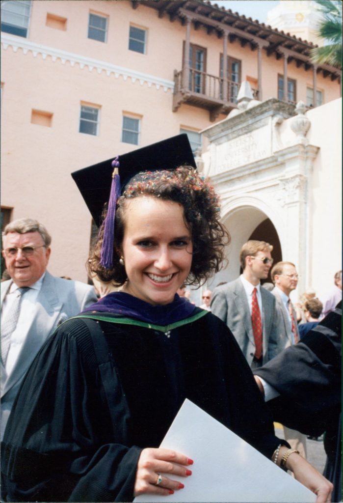 A woman wearing a cap and gown smiles at the camera after a graduation ceremony. There is confetti in her hair.