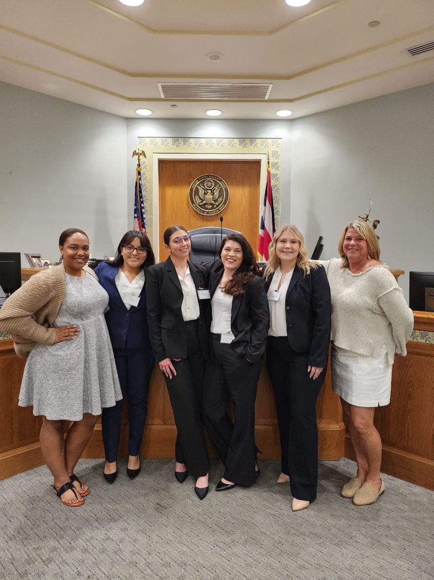 A group of female students dressed professionally poses in front of the dais in a practice courtroom.