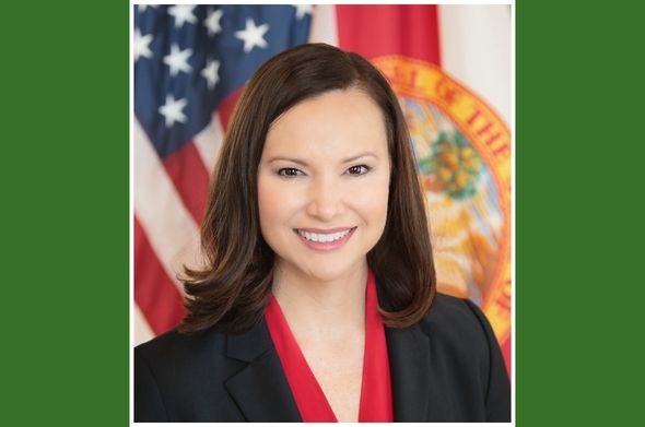 A woman with shoulder-length brown hair and brown eyes smiles for a headshot photo. The U.S. and Florida flags are in the background.