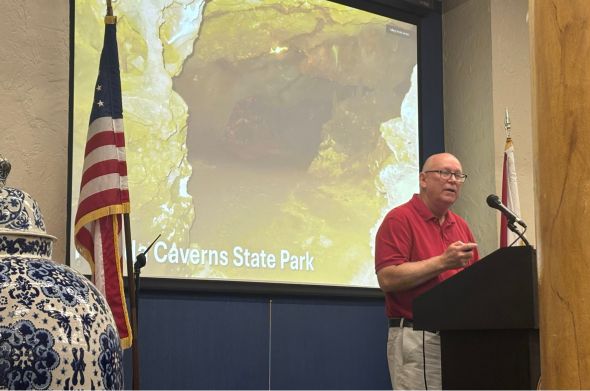 A man in a red shirt and glasses speaks at a podium. A picture of a large cavern is on the screen behind him.