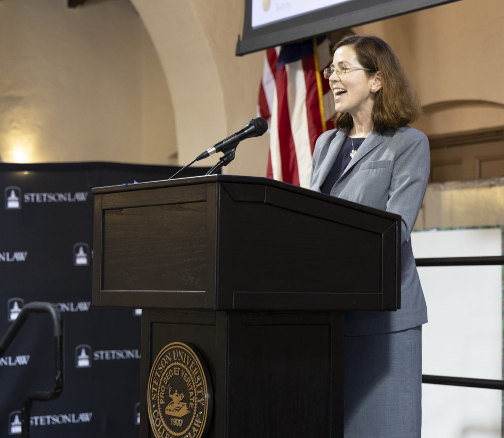 A woman with shoulder-length brown hair and glasses wearing a great suit and smiles as she speaks at a podium with the Stetson Law seal.