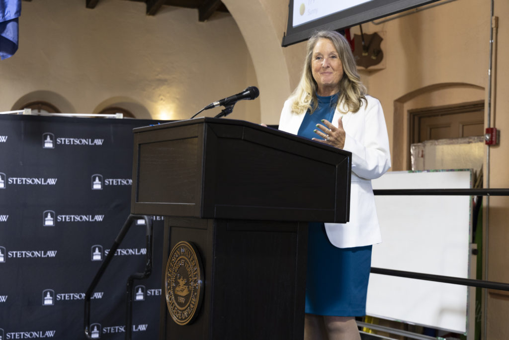 A woman in a white suit jacket speaks on a stage in Stetson Law's Great Hall.