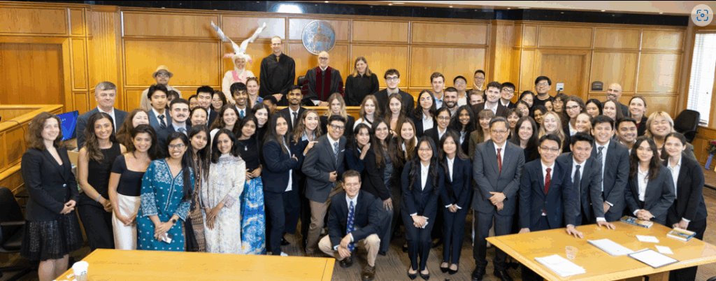 Dozens of students, faculty, and staff members stand in a practice courtroom. They are professionally dressed, except for a person who as dressed as a markhor, a species of goat that is the national animal of Iran.