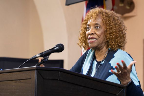 A woman with curly brown hair and a vibrant expression speaks at a podium.