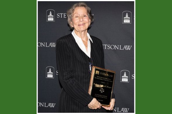 A woman with short hair smiles as she holds a rectangular award in front of a backdrop that has the words "STETSON LAW' printed on it.