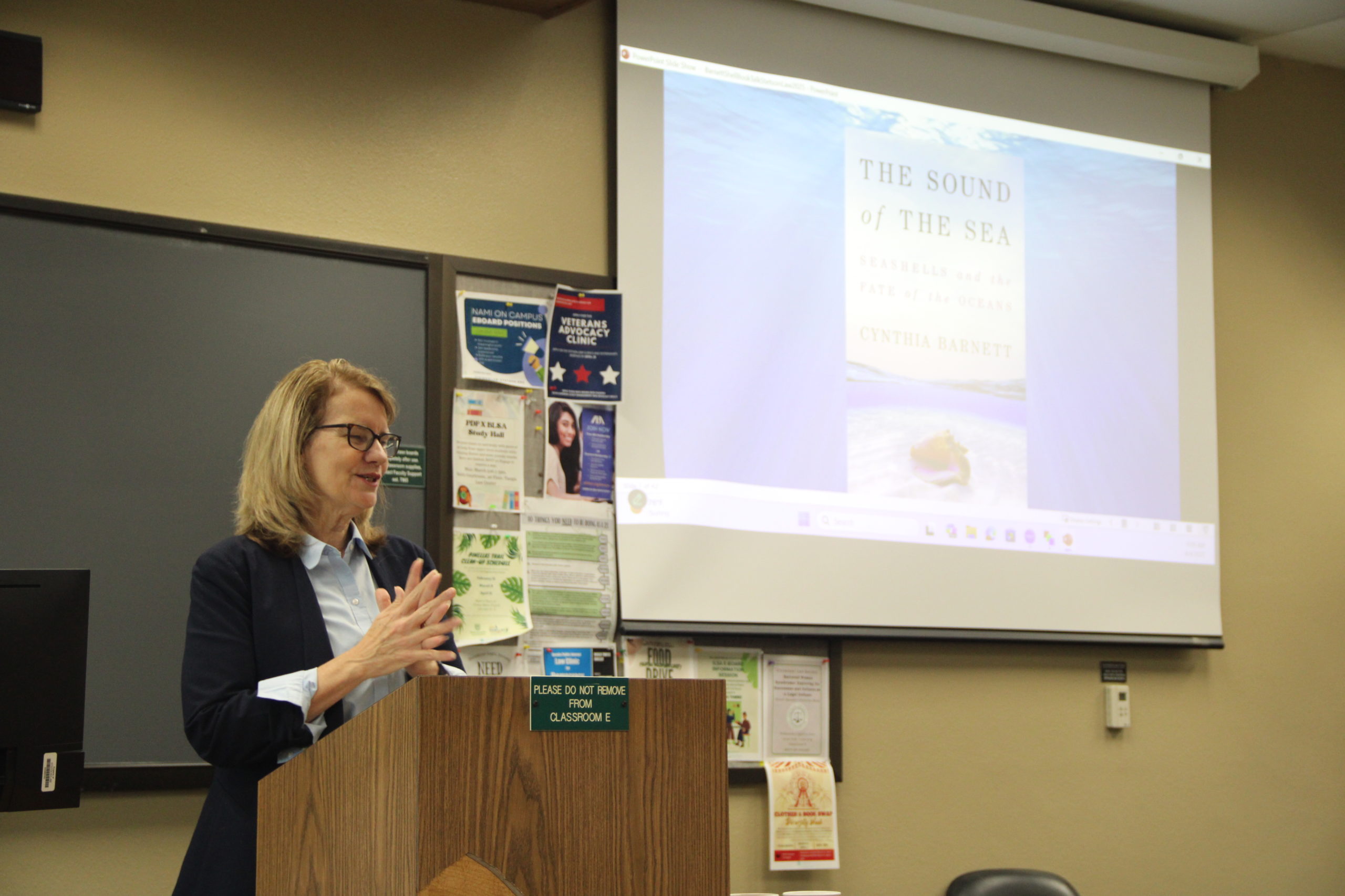 A woman speaks at the front of a classroom. a photo of her book, "Sound of the Sea," is projected on the wall behind her.