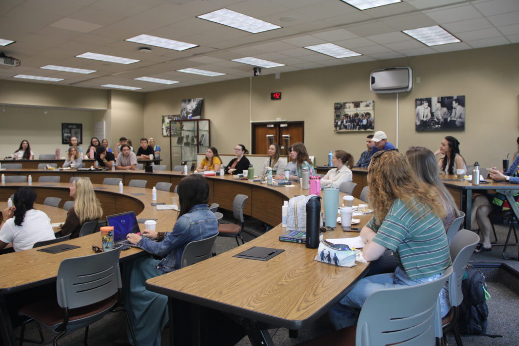 Students sit at several rows of semicircular classroom tables and engage in a discussion.