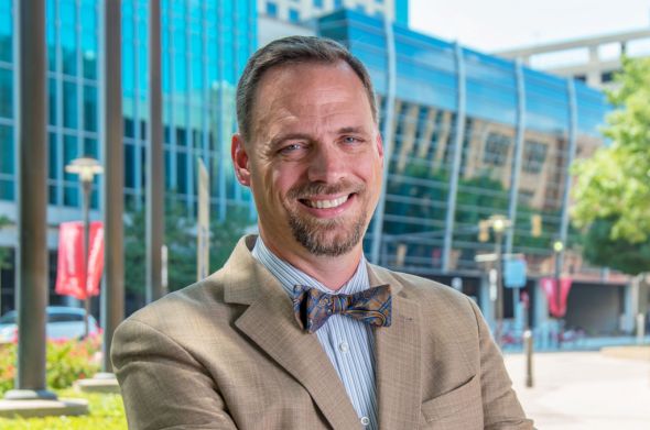 A man with blonde hair and a goatee in a khaki-colored jacket and bowtie smiles for the camera.